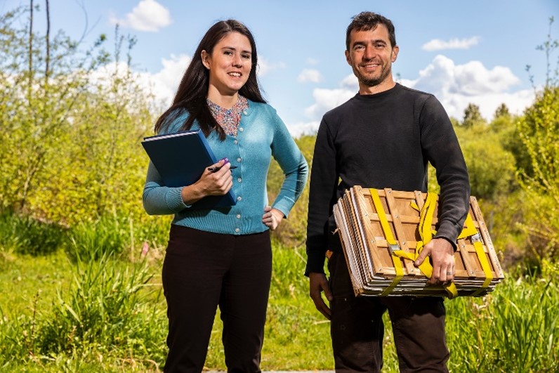 two UW employees at the Botanic Gardens stand in a sunny field, the woman holds a clipboard and the man a botanical press frame
